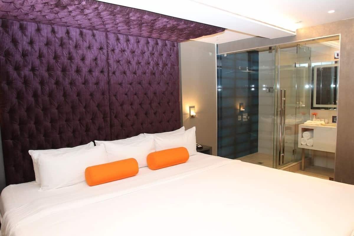 Een hotelkamer met een gestuikt hoofdeinde, een bed met witte lakens en twee oranje kussens, een douche met glazen wanden en een witte wastafel.