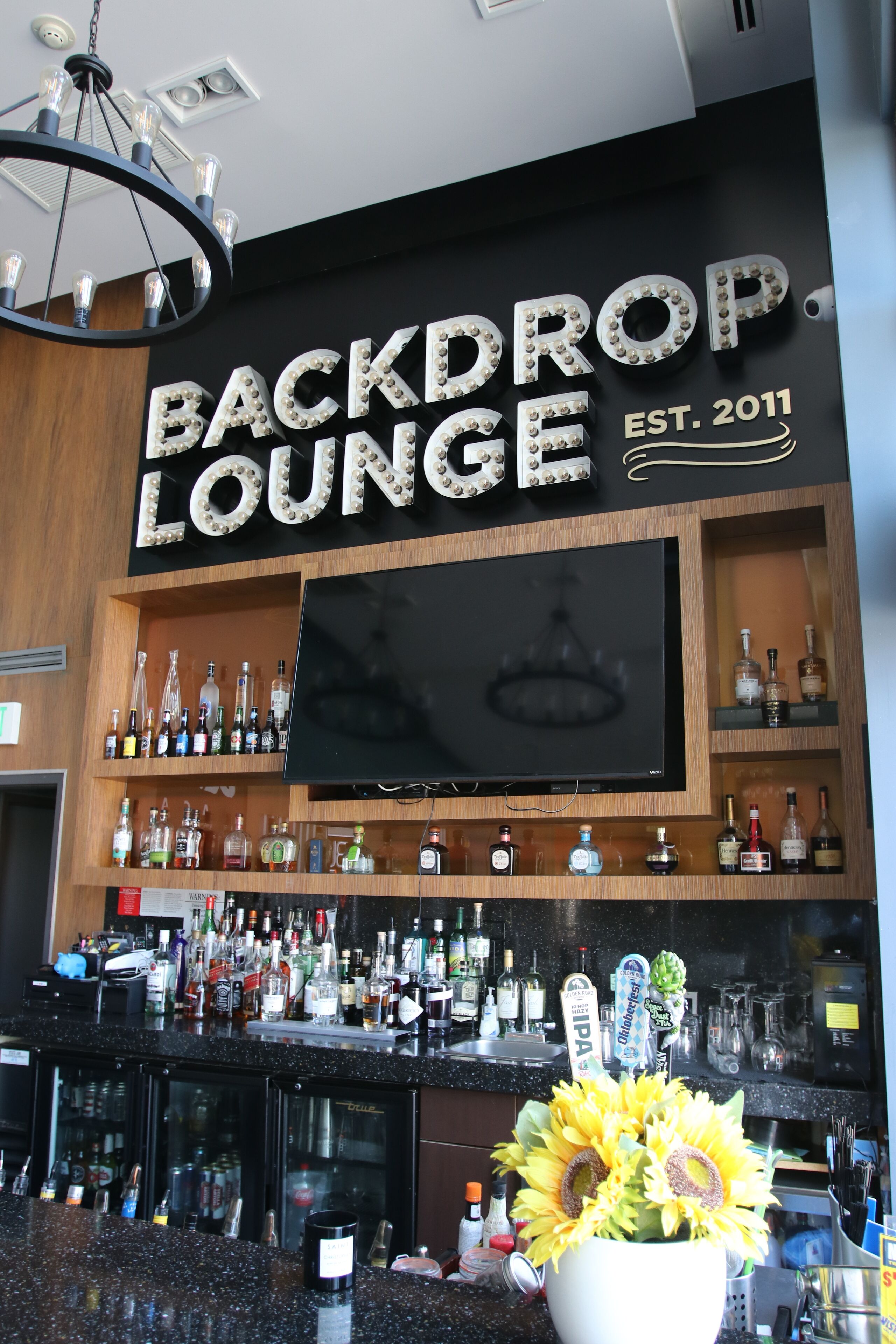 A bar area with a large sign reading "BACKDROP LOUNGE EST. 2011" and a TV mounted above the bar.