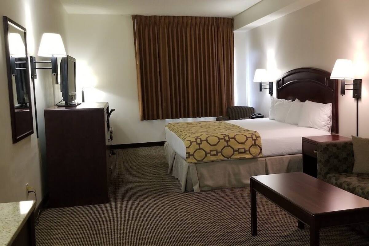 A hotel room with a bed, a desk, a chair, a lamp, and a large window with curtains.