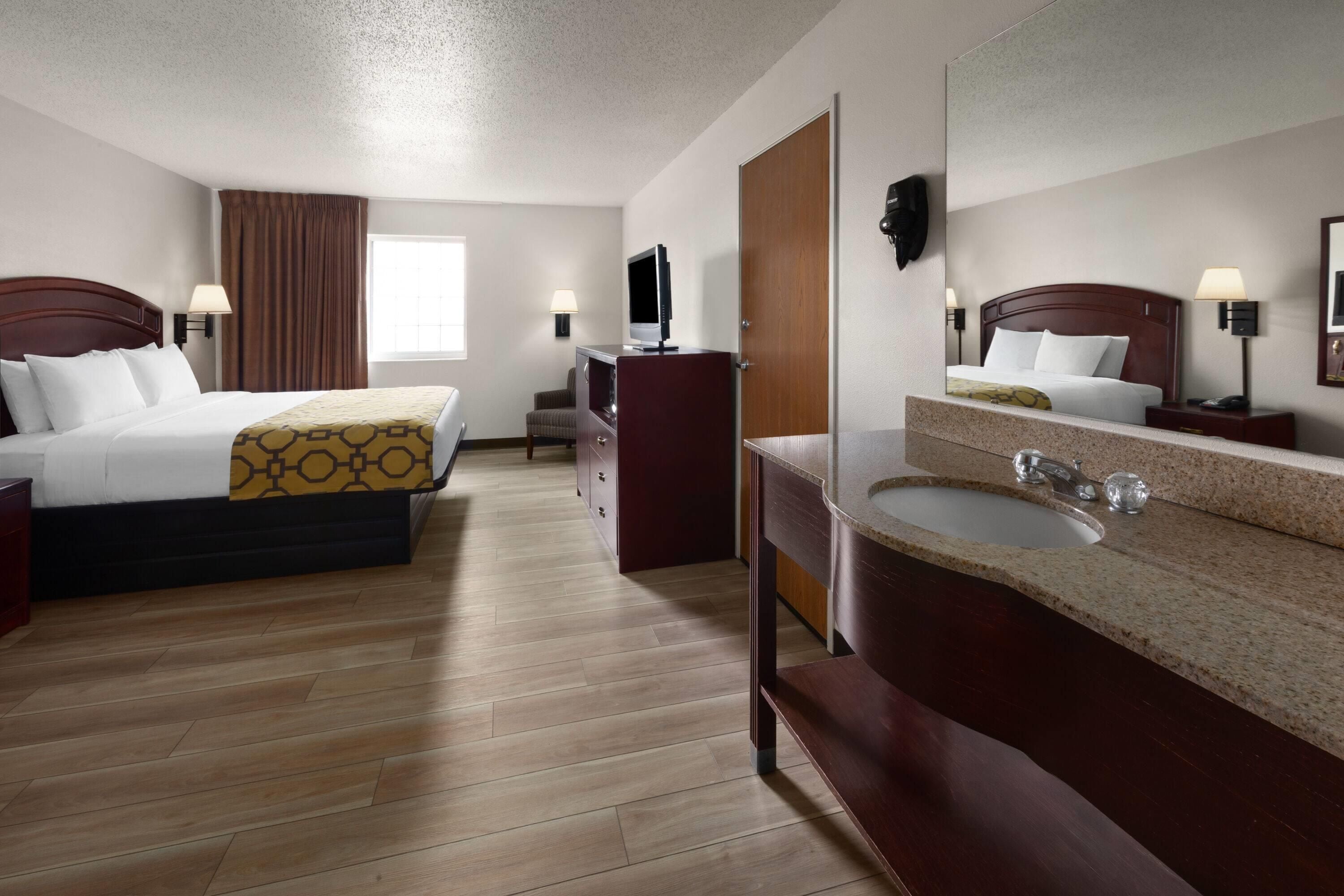 A hotel room with two beds, a desk, a TV, a mirror, and a bathroom with a sink and mirror.
