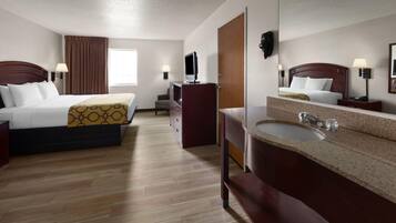 A hotel room with two beds, a desk, a TV, a mirror, and a bathroom with a sink and mirror.