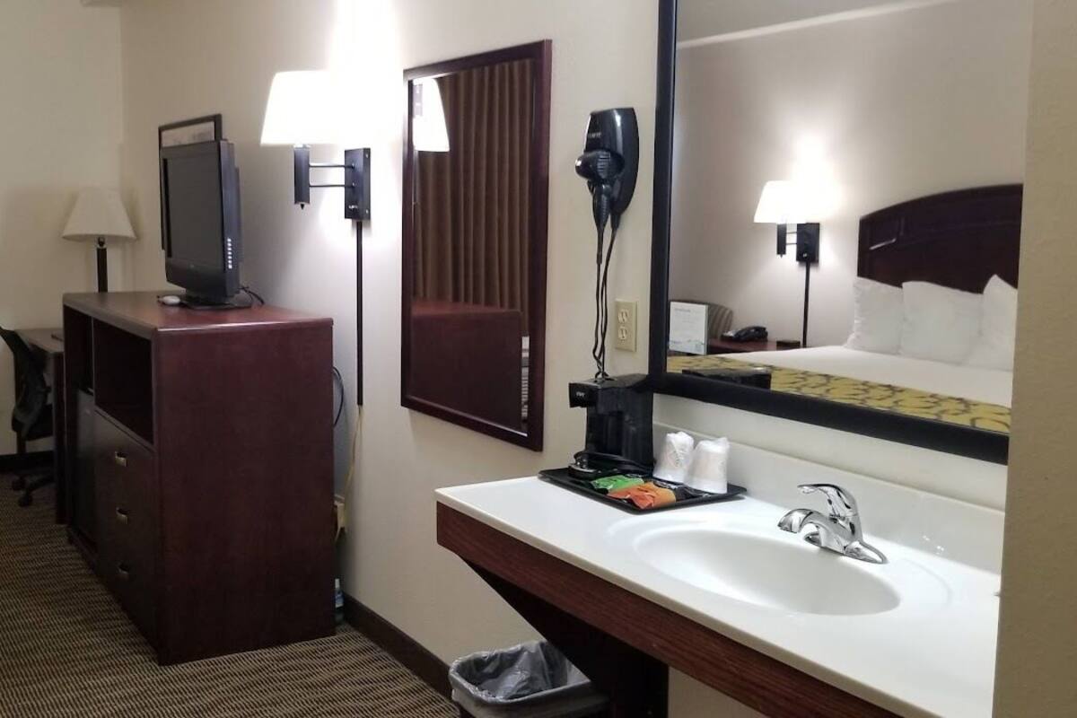 A hotel room with a bed, a desk with a computer, a mirror, a sink, and a hairdryer.