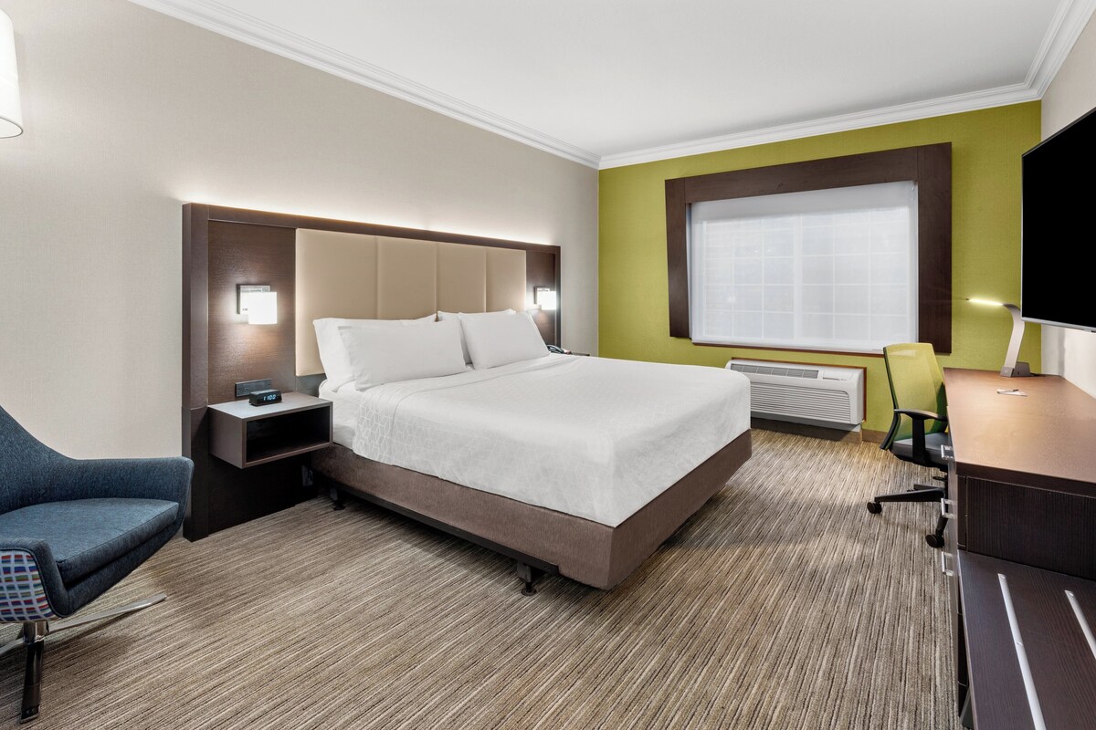 A hotel room with a bed, bedside table, chair, desk, and a large window.