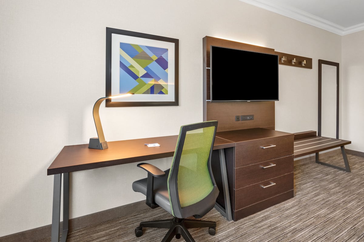 A hotel room with a desk, chair, TV, and artwork.