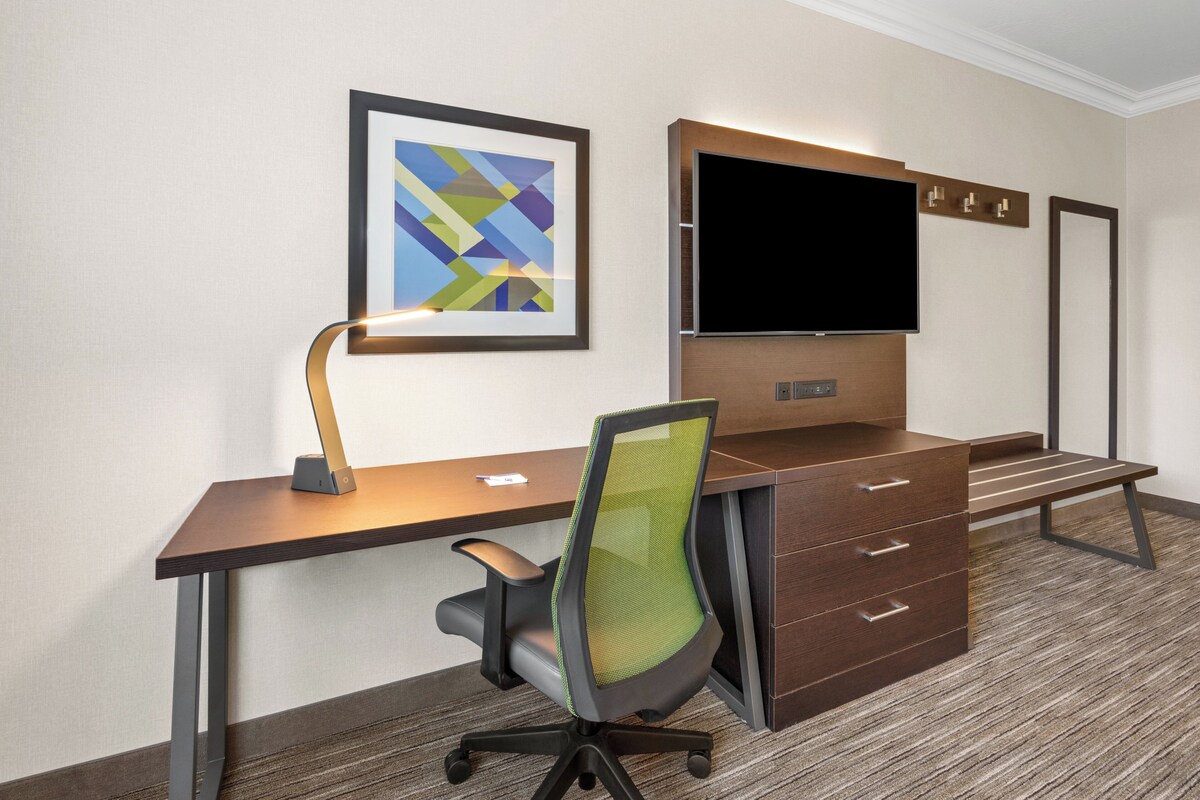 A hotel room with a desk, chair, TV, and artwork.