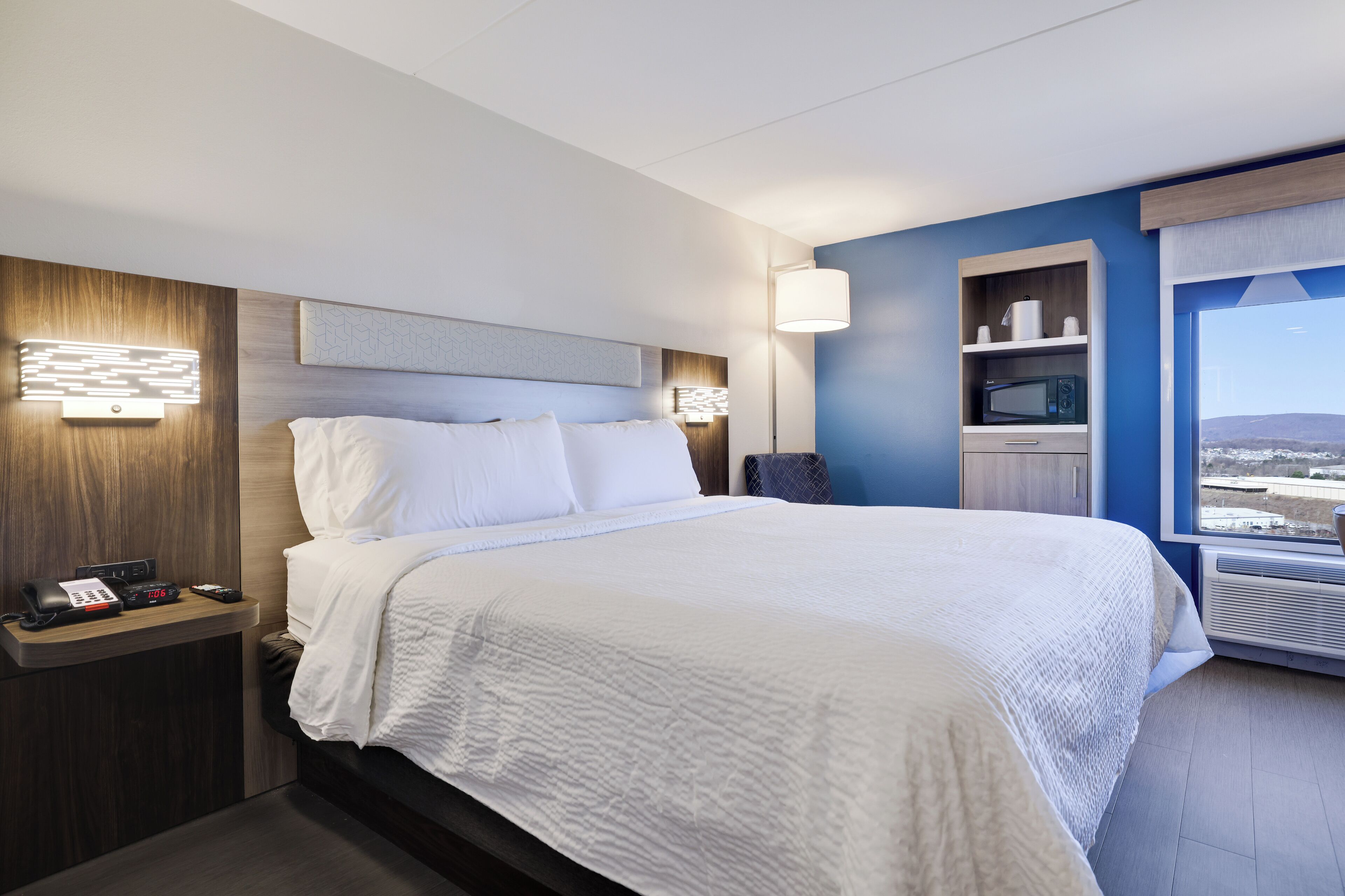 A hotel room with a large bed, bedside tables, a desk, a microwave, and a view of the city.