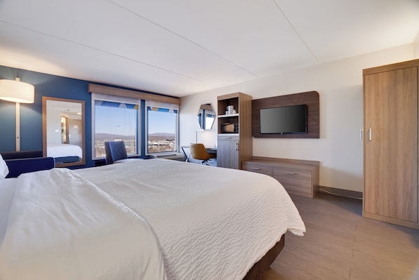 Quarto de hotel moderno com uma cama grande, uma escrivaninha, uma cadeira, um guarda-roupa, uma televisão e vista para a cidade.
