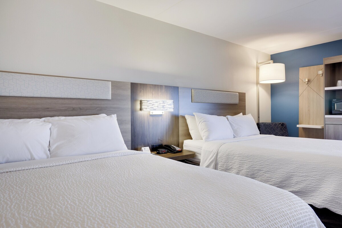Quarto de hotel com duas camas, uma mesa de cabeceira com telefone e luminária, e uma prateleira na parede.