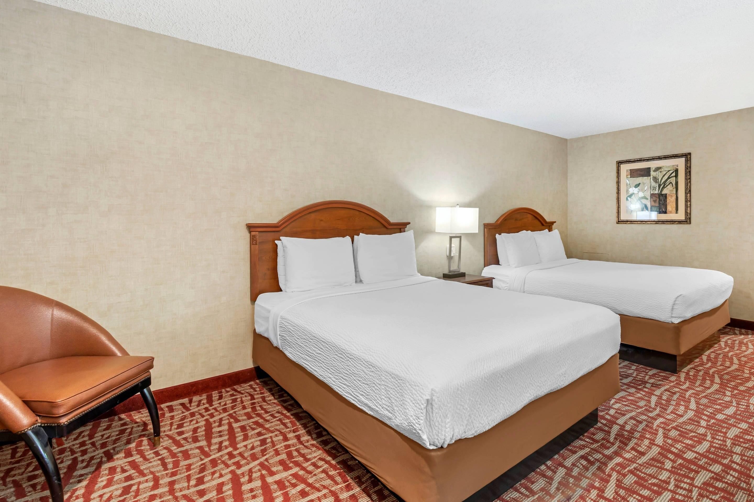 A hotel room with two beds, a chair, a lamp, and a painting on the wall.