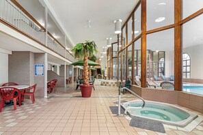 An indoor swimming pool area with a hot tub, red chairs, and a palm tree.