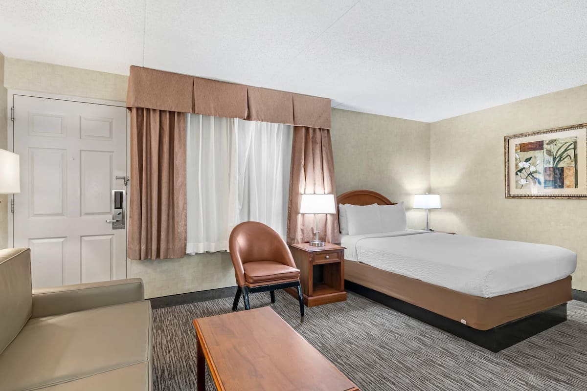 A hotel room with a bed, a chair, a desk, a lamp, and a painting on the wall.