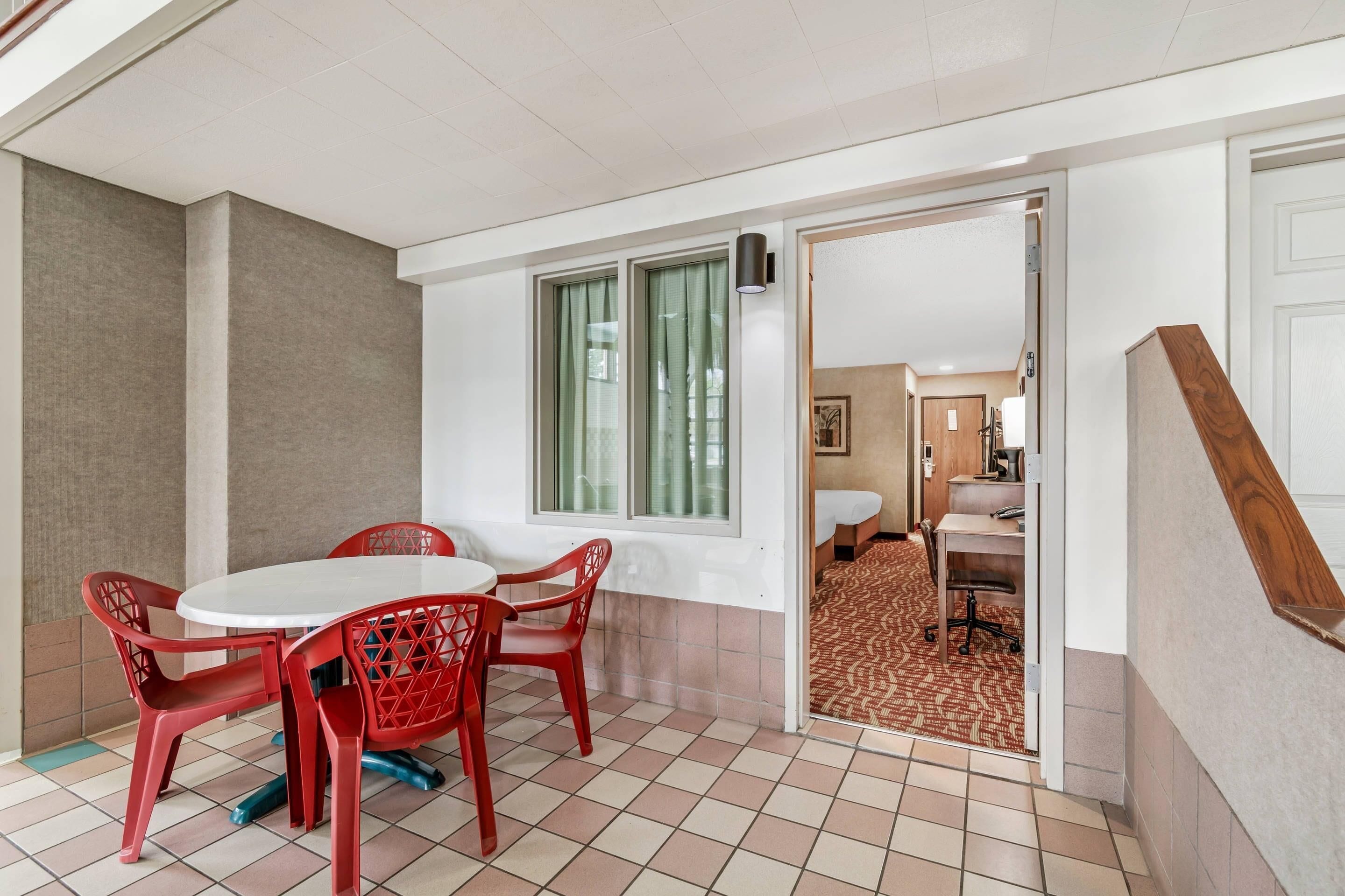 Una habitación con una mesa redonda blanca y sillas rojas, una ventana con cortinas, una puerta que da a otra habitación y un suelo de baldosas.