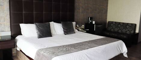 A hotel room with a large bed, a dark headboard, a bedside table with a lamp, and a patterned sofa.