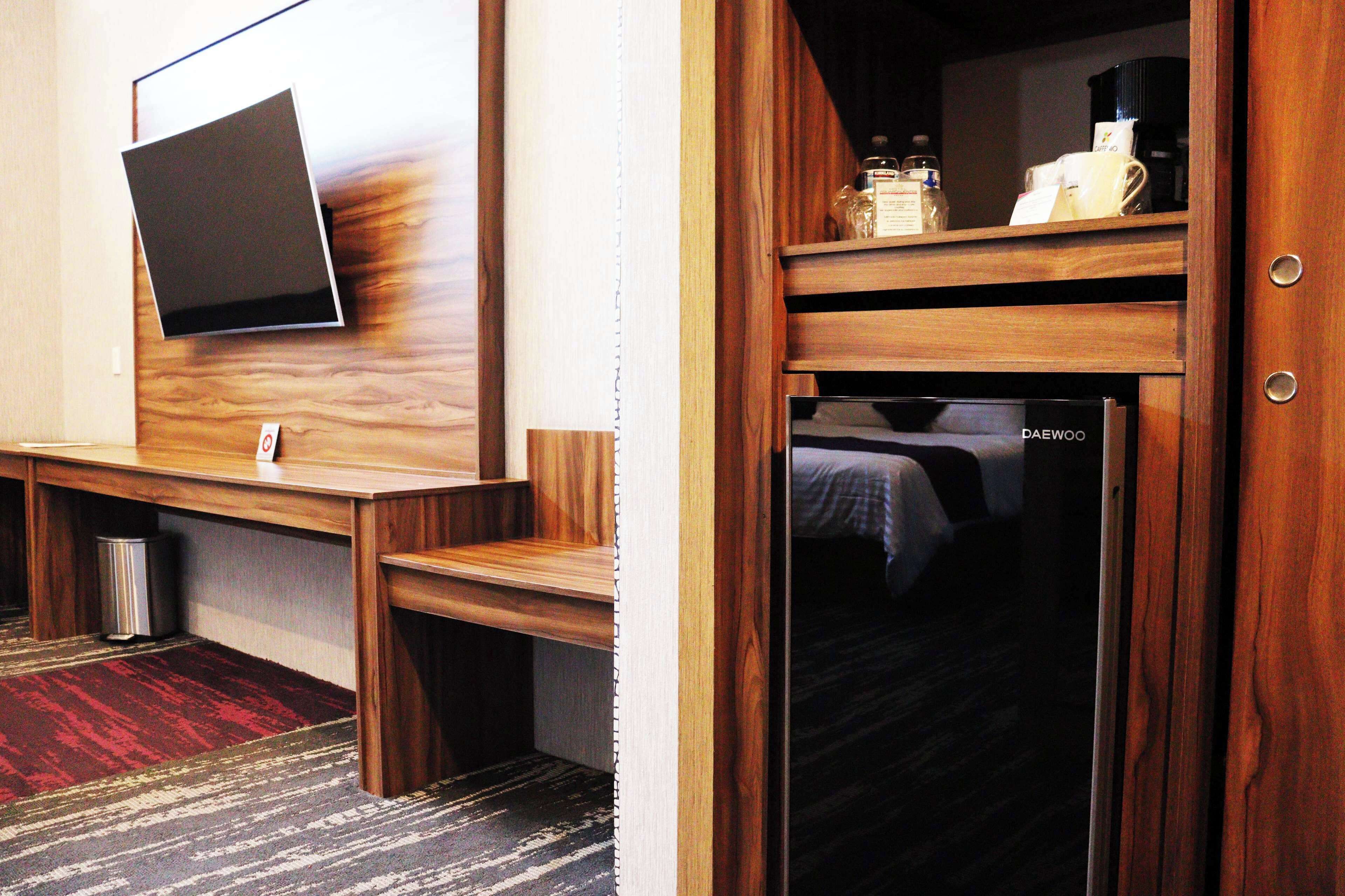 Quarto de hotel com televisão de tela plana na parede, escrivaninha de madeira, cama com cabeceira preta e minibar.