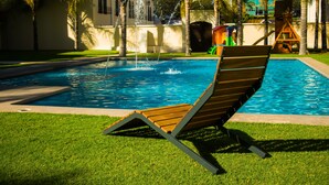 A pool with a wooden lounge chair, palm trees, and a water fountain.