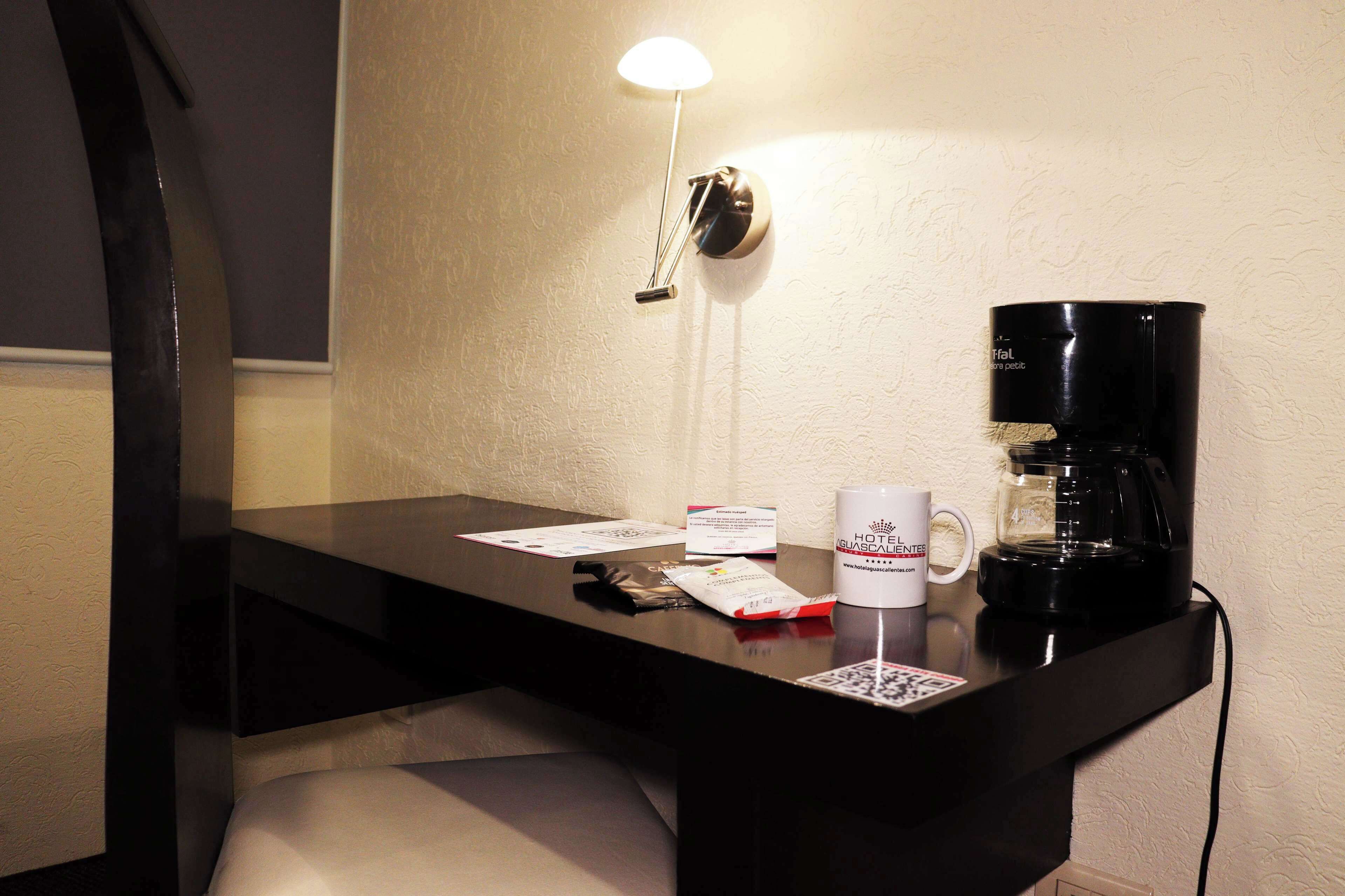 A hotel room desk with a coffee maker, a mug, a lamp, and some papers.