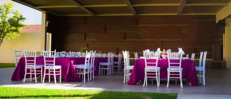 Espaço para eventos ao ar livre com mesas montadas para convidados, cadeiras brancas e toalhas de mesa roxas.