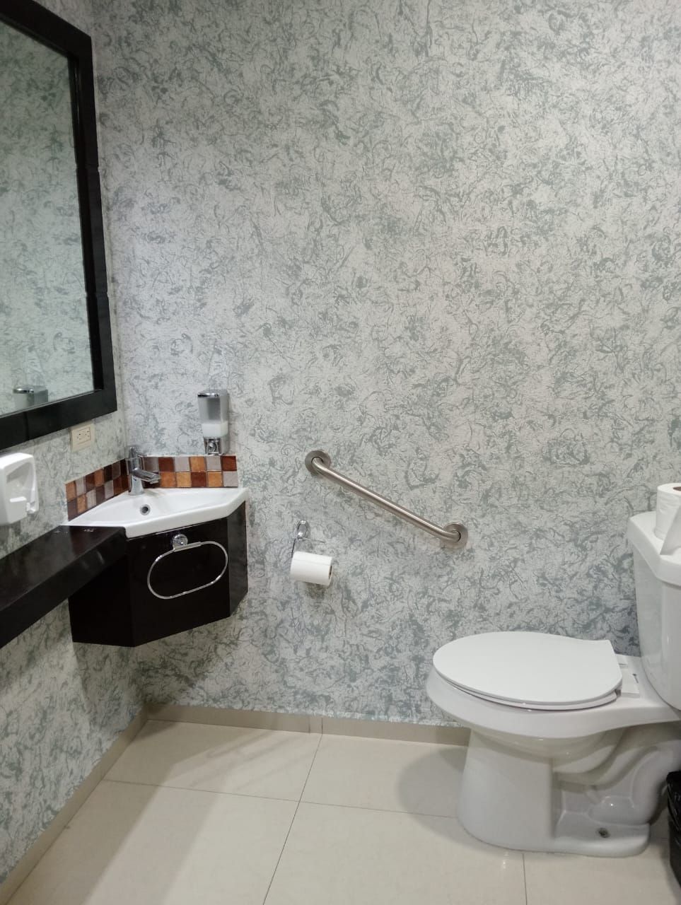 Un baño con pared de diseño marmolizado, un lavamanos negro con espejo, un inodoro y un pasamanos.