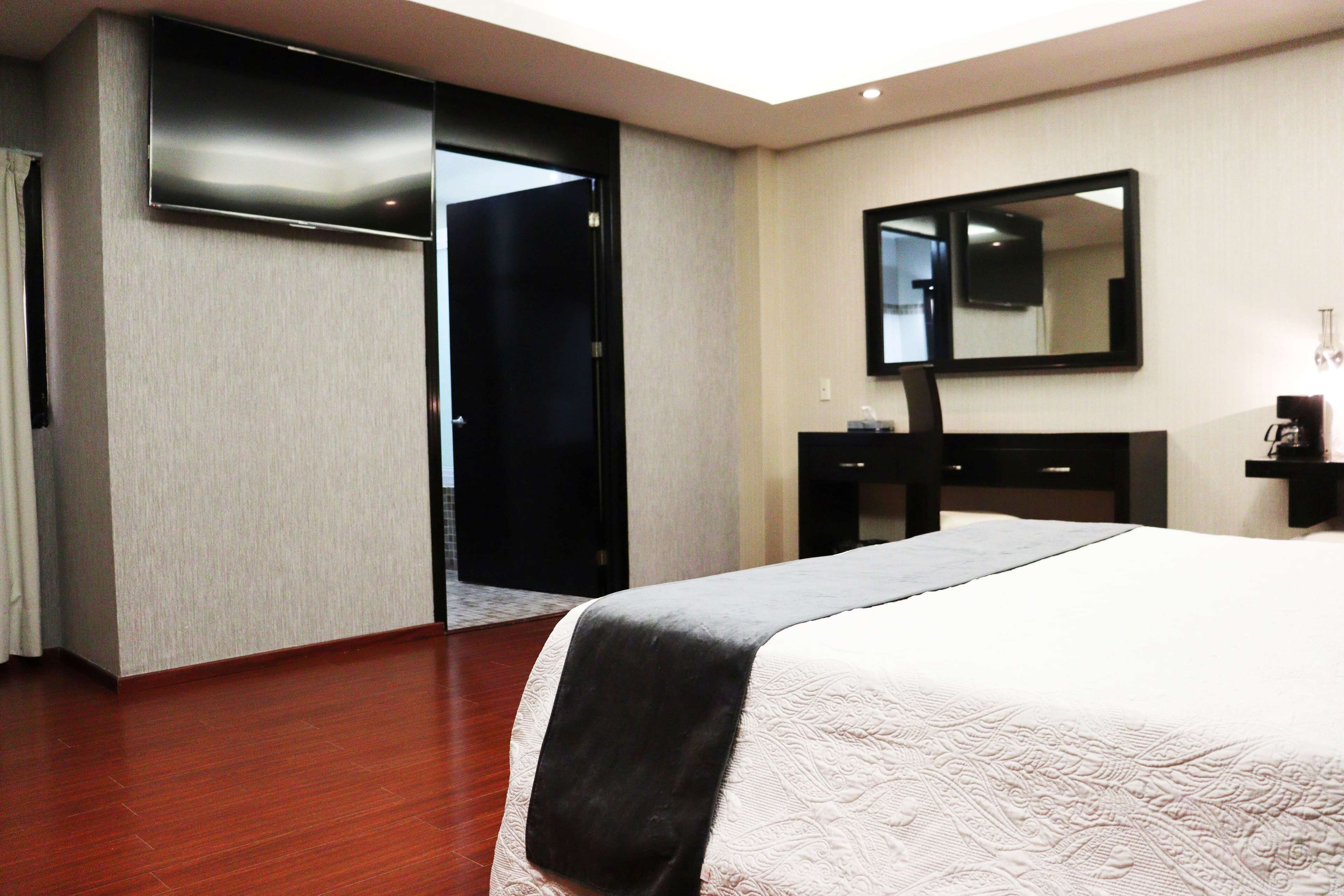 A hotel room with a bed, a flat-screen TV, a mirror, a dresser, and a door leading to another room.