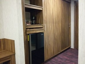 A room with a wooden wardrobe, a built-in refrigerator, and a small wooden shelf with items on it.