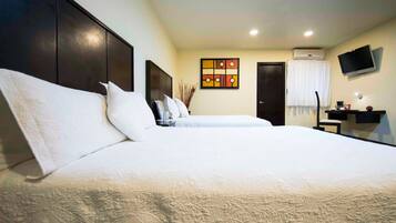 A hotel room with a large bed, a desk with a chair, a flat-screen TV, and an air conditioning unit.