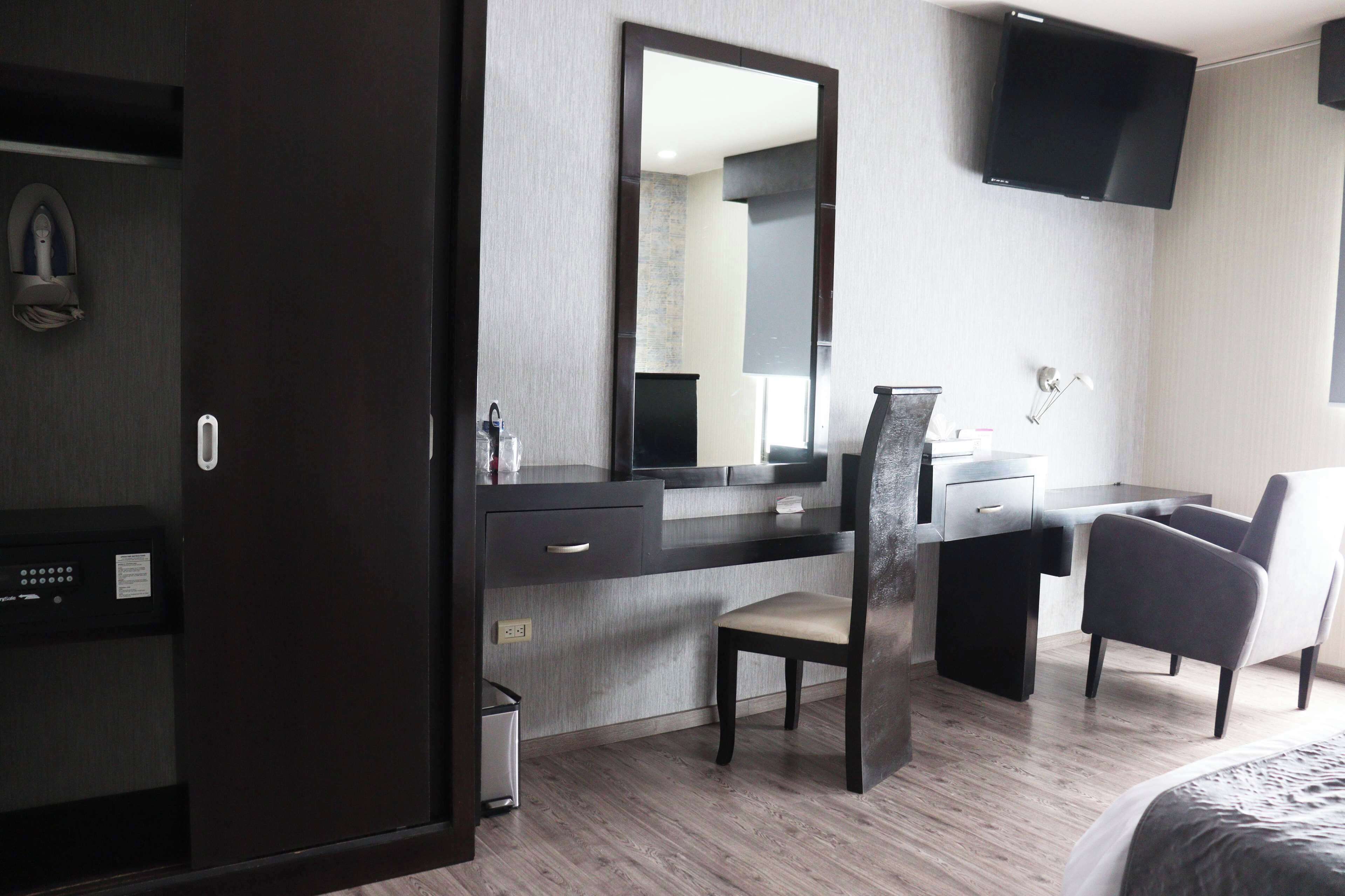 A modern hotel room with a dark wood desk, a large mirror, a flat-screen TV, and a comfortable chair.