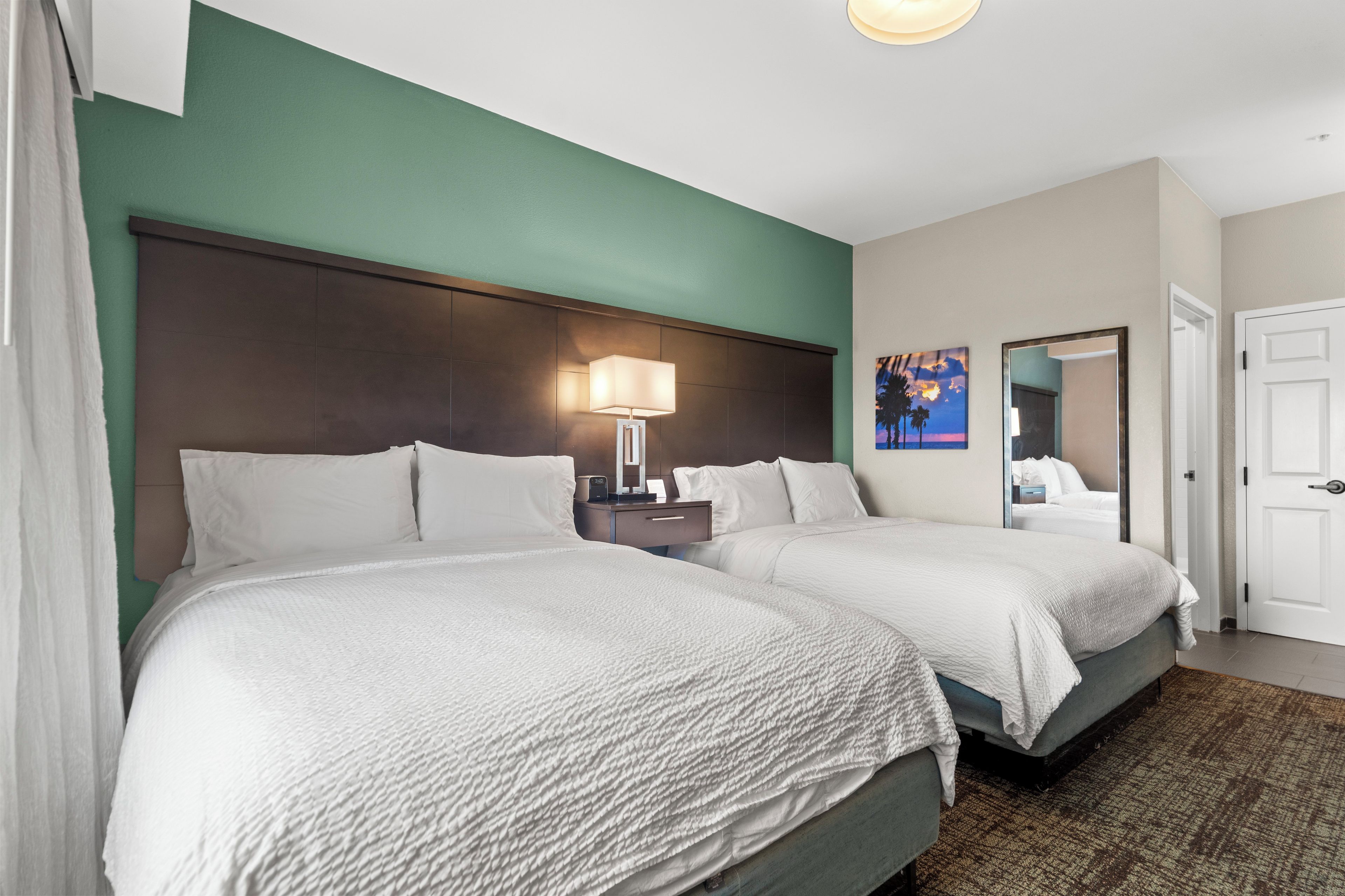 A hotel room with two beds, a nightstand with a lamp, a painting on the wall, and a door.