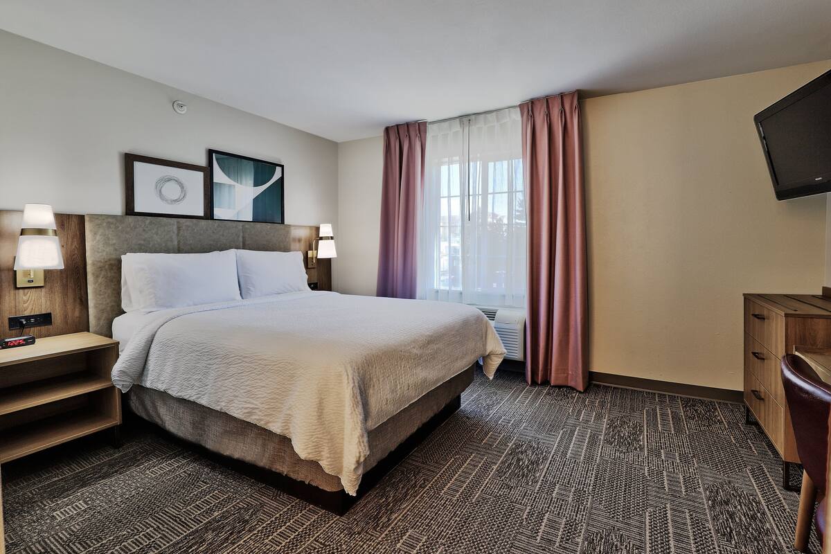 Une chambre d’hôtel avec un lit, des tables de chevet avec des lampes, un téléviseur à écran plat, une grande fenêtre avec des rideaux et une moquette à motifs.