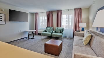 A hotel room with a flat-screen TV, two green armchairs, a beige sofa, a small round table, and a brown ottoman.