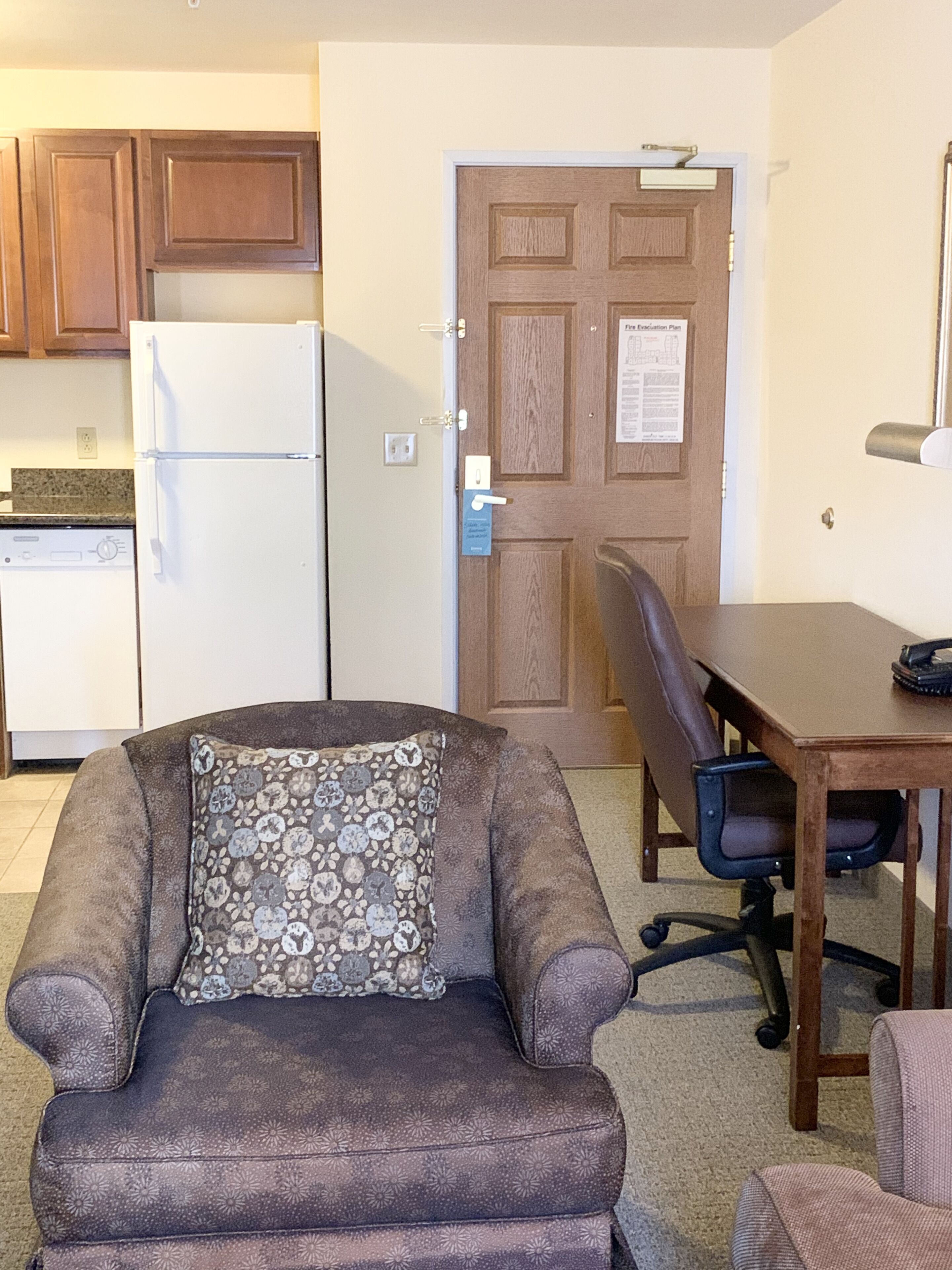 A compact living space with a kitchenette, a brown sofa with a patterned pillow, a wooden dining table, and a black office chair.