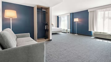 A modern hotel room with a grey sofa, a bed, a walk-in closet, and a large window with curtains.