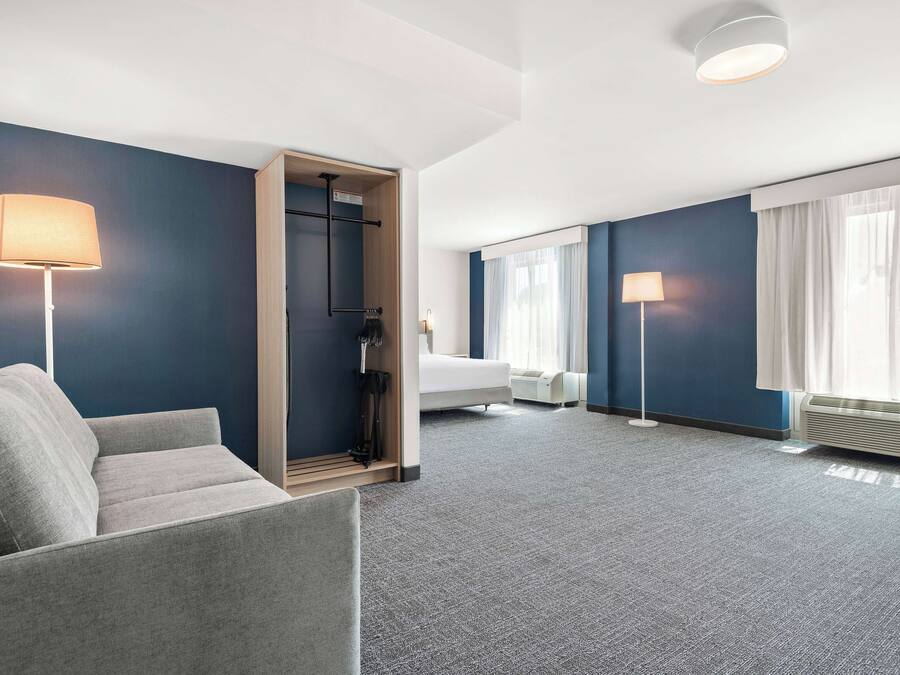 A modern hotel room with a grey sofa, a bed, a walk-in closet, and a large window with curtains.