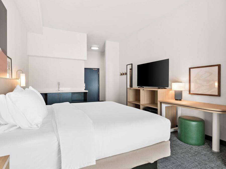 A hotel room with a bed, a desk, a television, and a green stool.