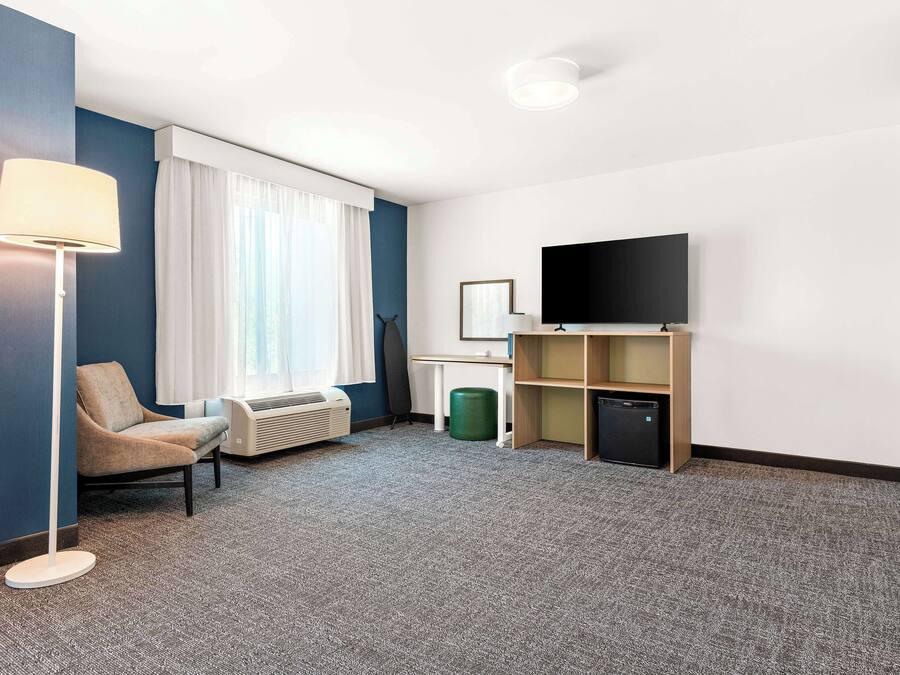 A hotel room with a flat-screen TV, a chair, a desk, a lamp, and a window with curtains.
