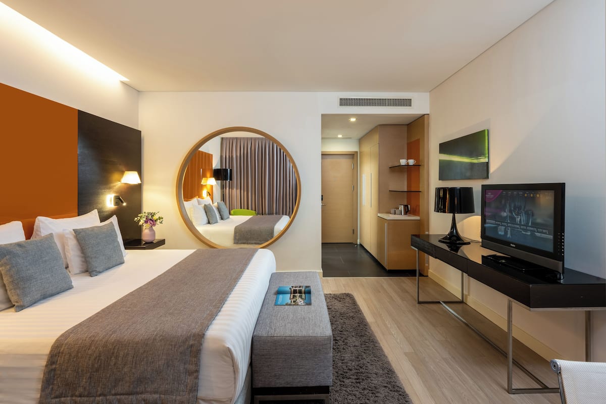 Een moderne hotelkamer met een groot bed, een bureau met een flatscreen televisie, een ronde spiegel en een apart zithoekje met een fauteuil.