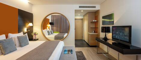 A modern hotel room with a large bed, a desk with a flat-screen TV, a round mirror, and a separate seating area with a chair.