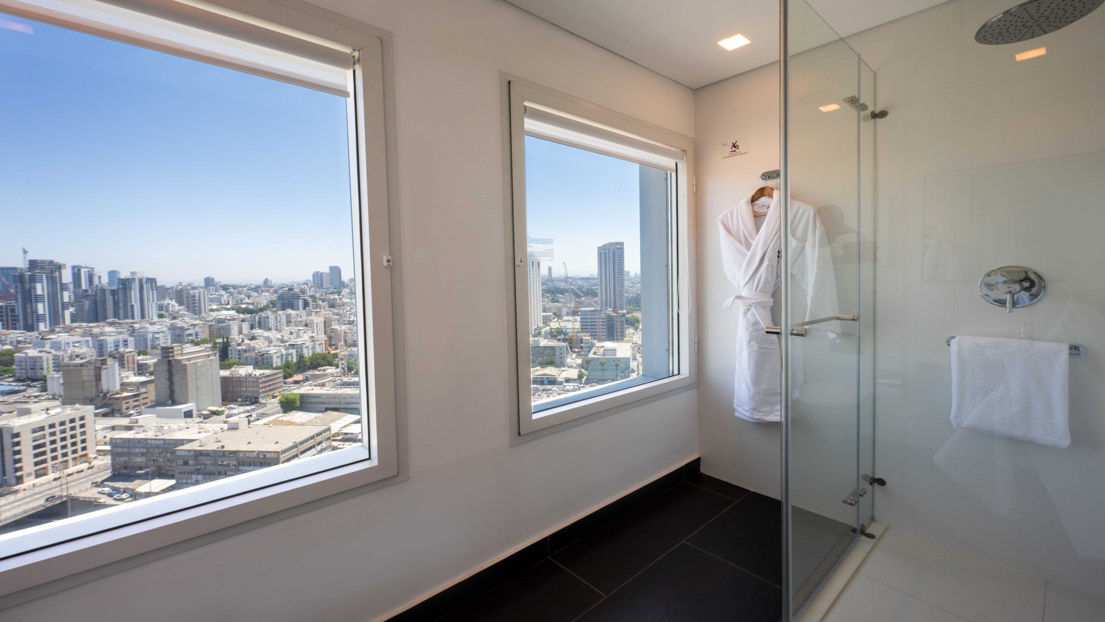 A modern bathroom with a glass shower, a large window offering a city view, and a white towel hanging on the wall.