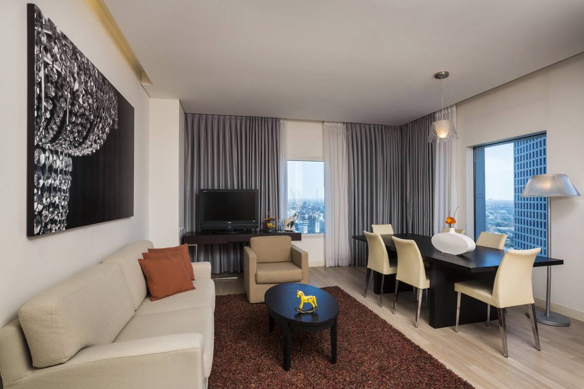 Een moderne hotelkamer met een bank, een eettafel met stoelen, een televisie en een groot raam met uitzicht op de stad.