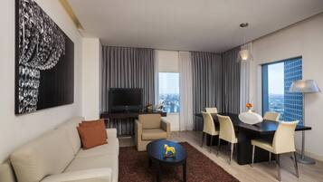 Una habitación de hotel moderna con sofá, una mesa de comedor con sillas, televisión y un ventanal con vistas a la ciudad.