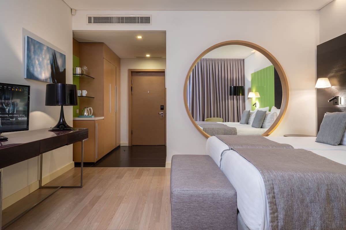 Een moderne hotelkamer met een bed, een bureau met een computer, een spiegel en uitzicht op de kamer via een ronde spiegel.