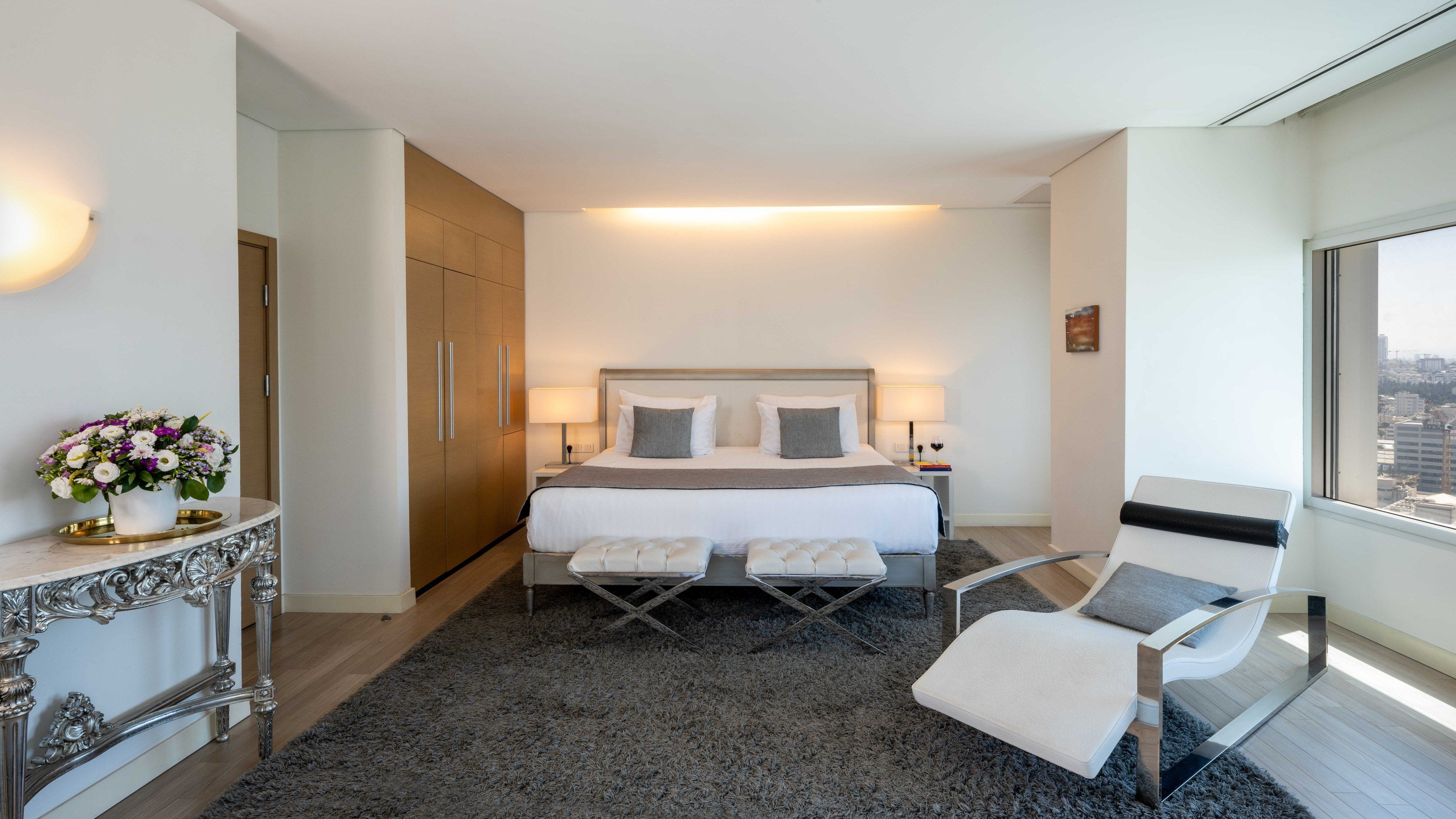 Habitación de hotel con cama, mesitas de noche, una silla, un jarrón decorativo y vista a la ciudad a través de una ventana.