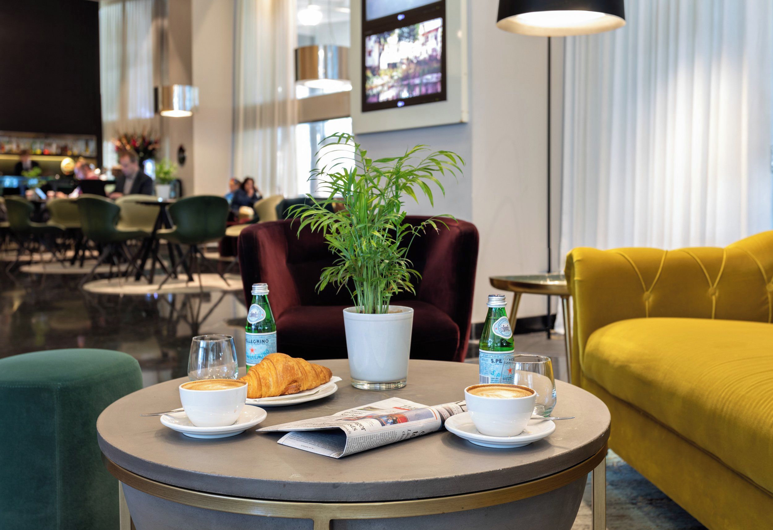 Ein runder Tisch mit einer Pflanze, einem Croissant und zwei Kaffeetassen, platziert in einem modernen Innenraum mit Sitzgelegenheiten und einem Fernsehbild im Hintergrund.