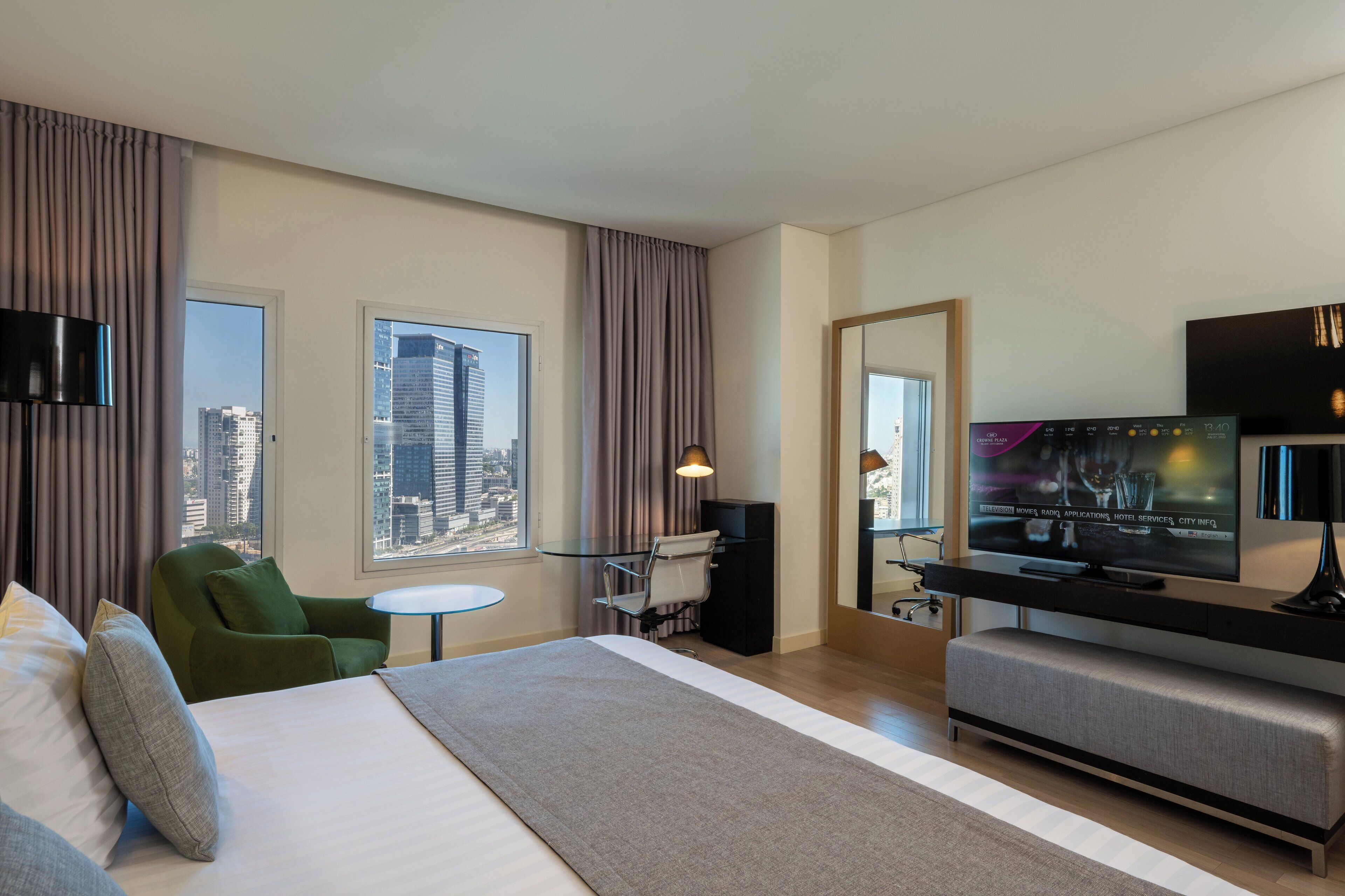 A modern hotel room with a large bed, a desk, a television, and a view of the cityscape.