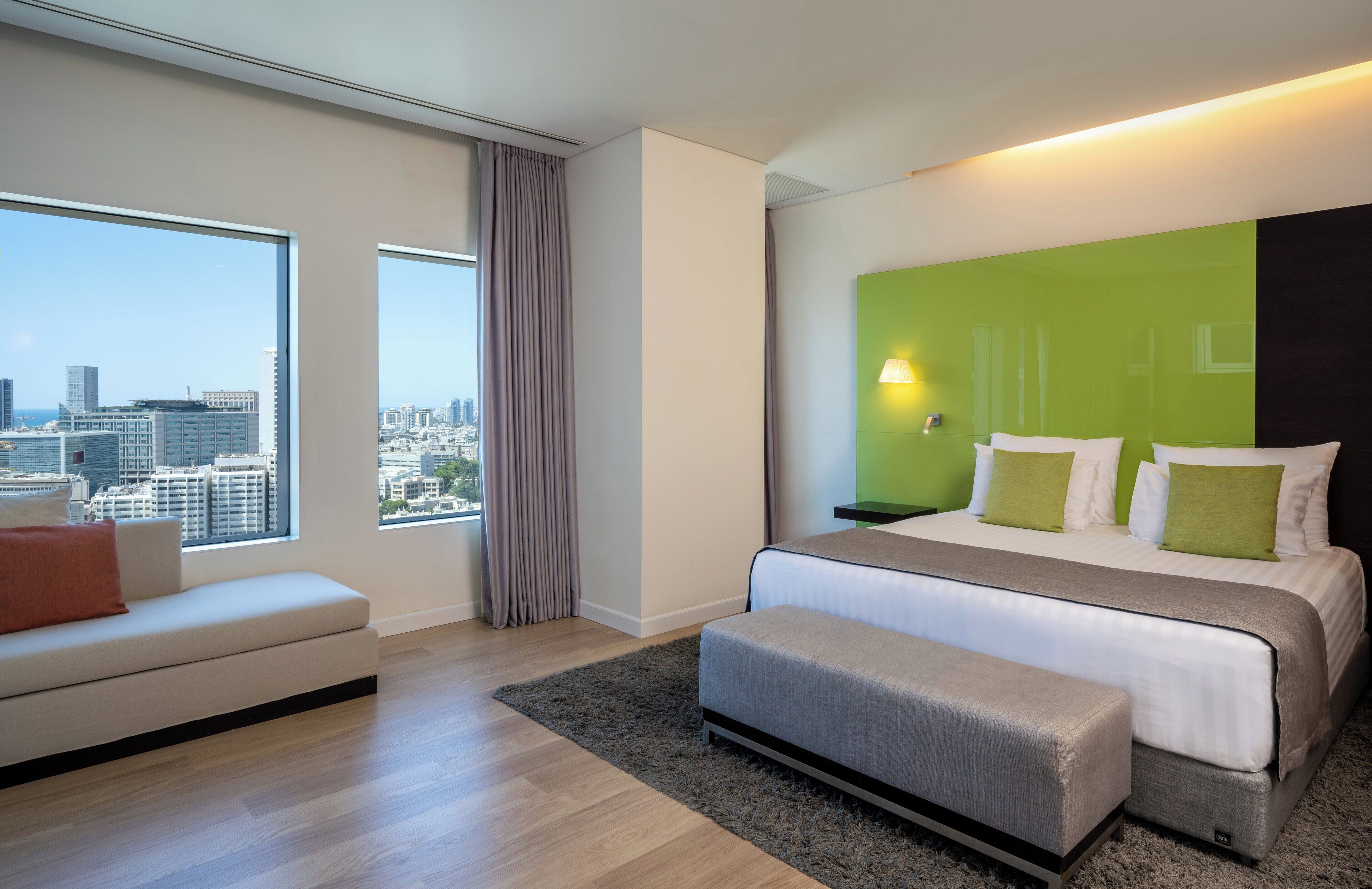 A modern hotel room with a large bed, a sofa, and a view of the cityscape.