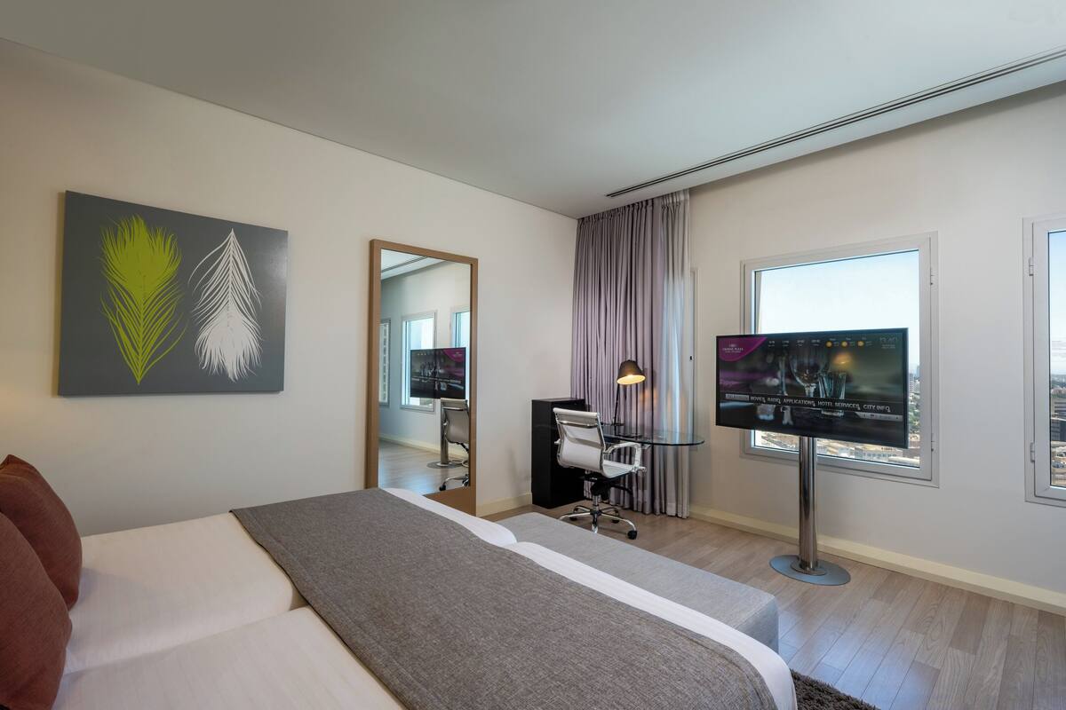 Een moderne hotelkamer met een groot bed, een bureau met computer, een flatscreen televisie en een groot raam met gordijnen.