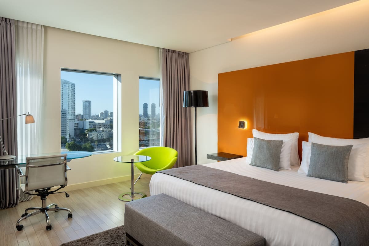 Een moderne hotelkamer met een groot bed, een bureau, een felgroene stoel en uitzicht op de stad door grote ramen.