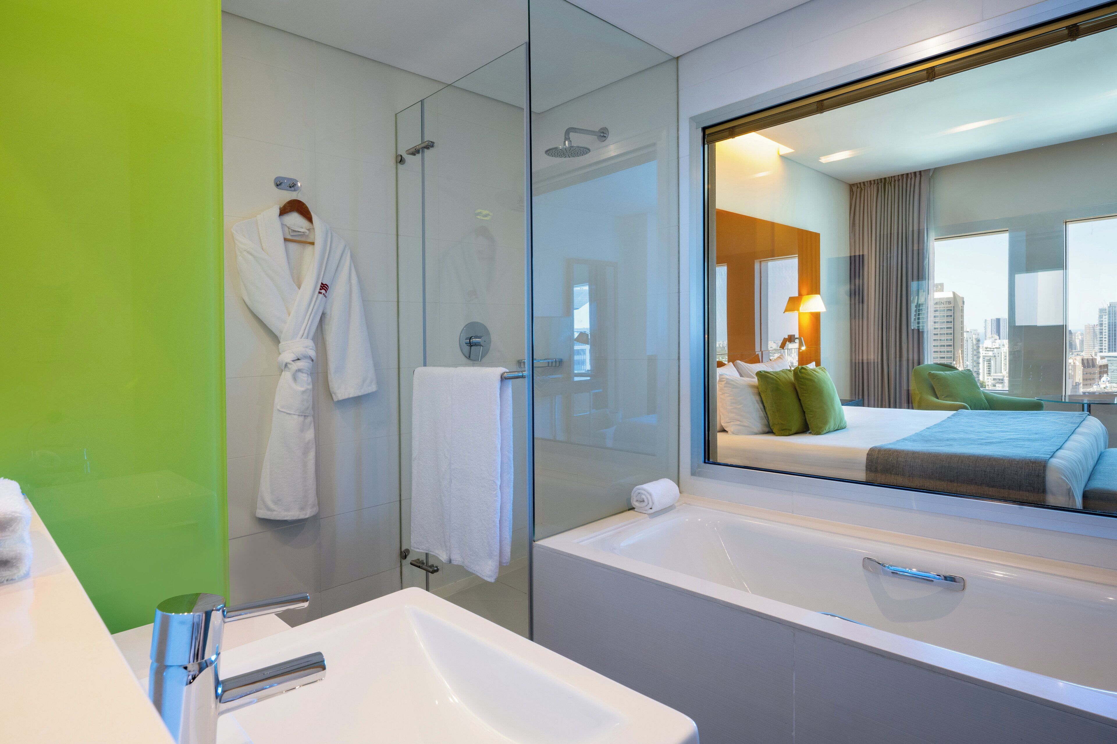 Une salle de bain d’hôtel moderne dotée d’une douche à parois de verre, d’une grande baignoire et d’un miroir qui reflète la vue sur la ville.