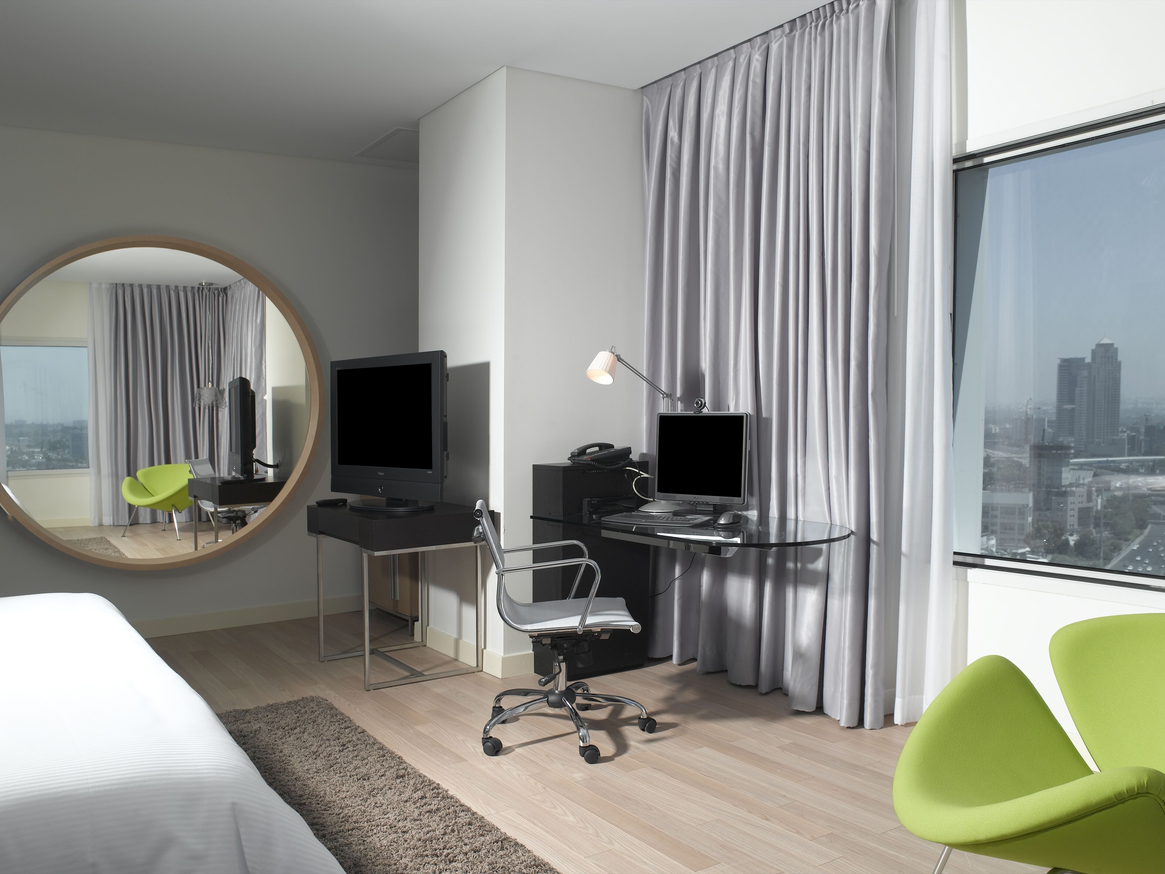 Una habitación de hotel moderna con una cama grande, un escritorio con una computadora, una silla, un televisor y un ventanal con vista a la ciudad.