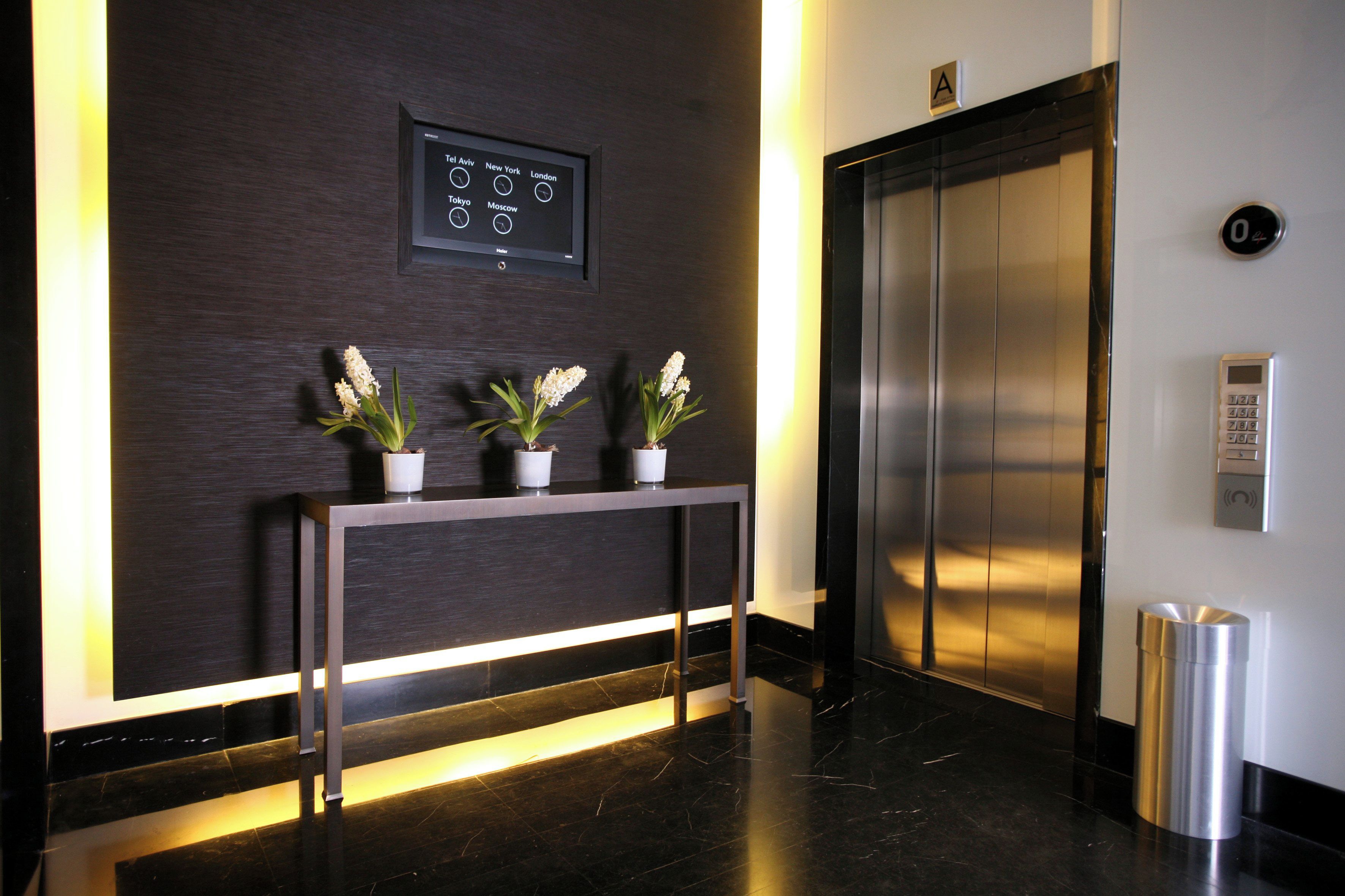 Uma área de recepção com um relógio digital exibindo horários em várias cidades, um balcão de atendimento com plantas em vasos e um elevador.