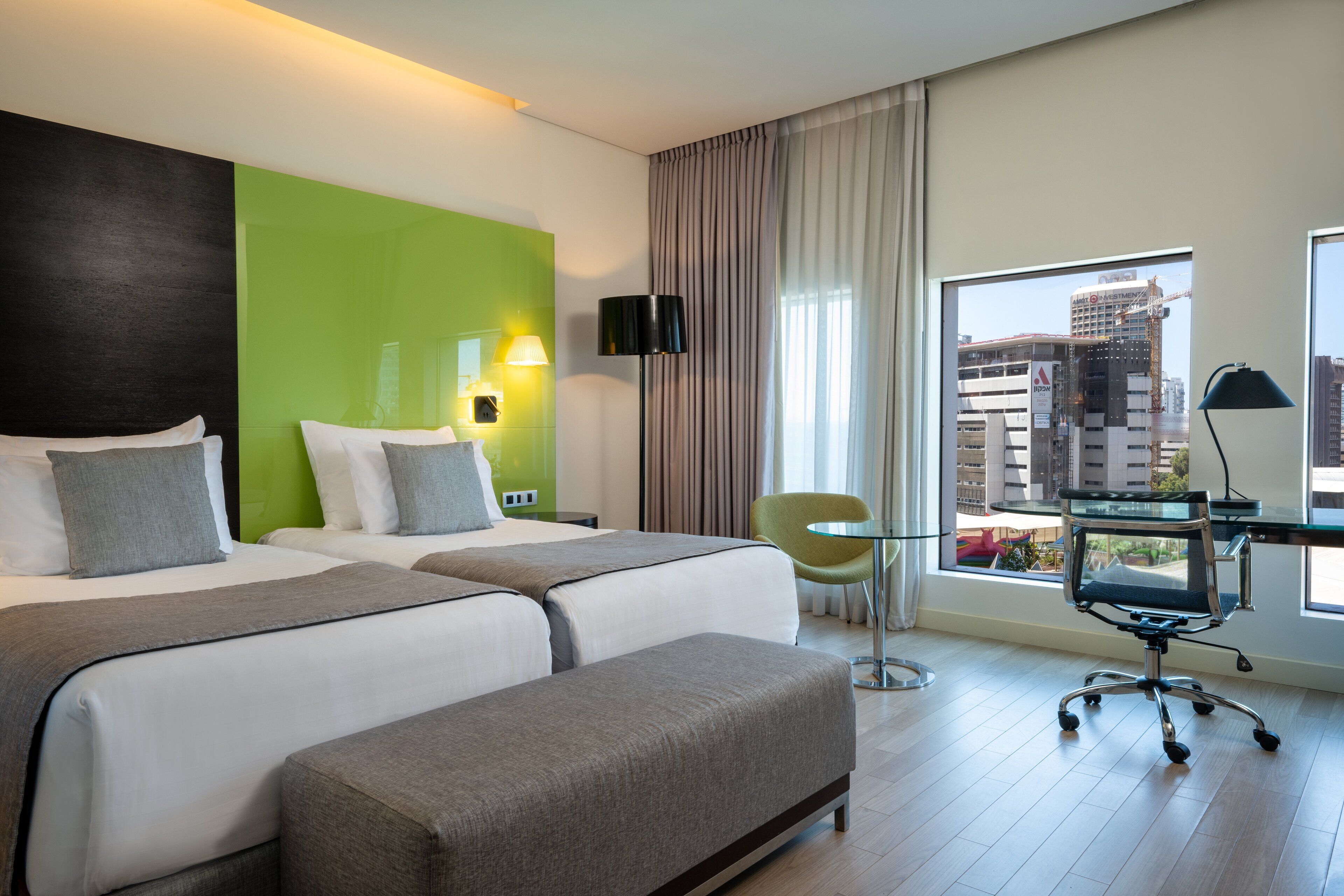 A hotel room with two beds, a desk with a chair, a green chair, and a view of the city.